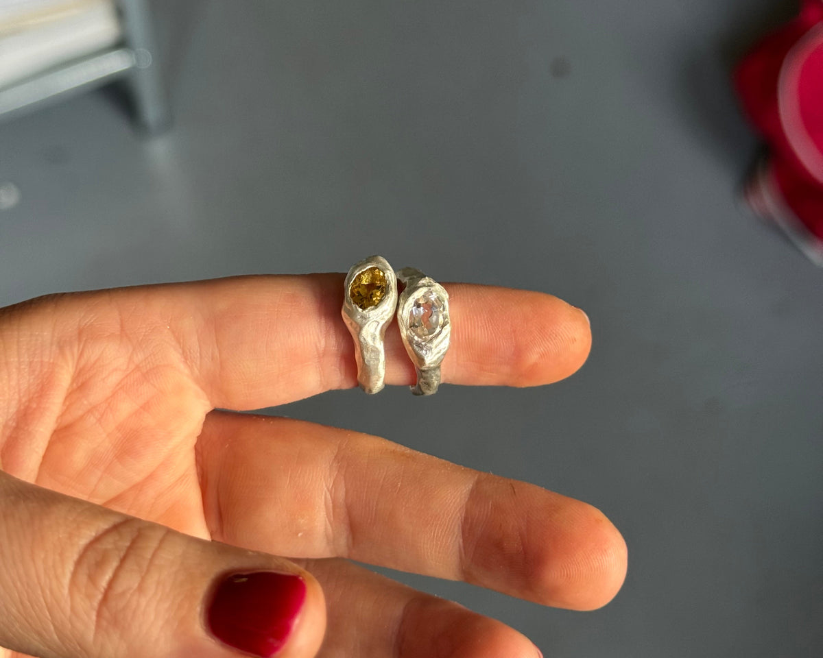 A hand with red nail polish holds two small rings against a gray indoor background. One ring is silver with a yellow gemstone, and the other is silver with a clear gemstone.