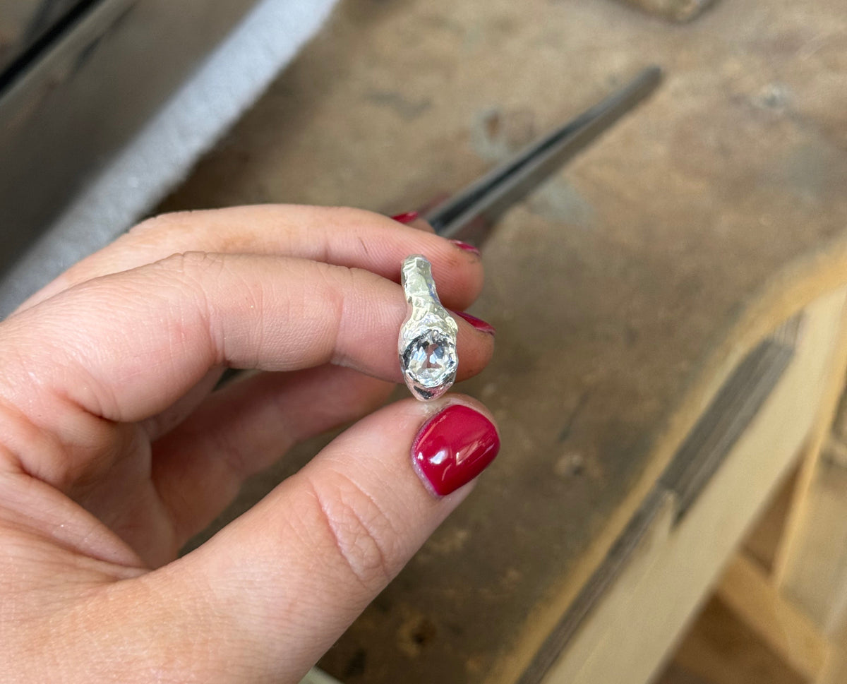 A hand holds an organic sterling silver ring with a clear round white topaz gemstone over a wooden workbench.
