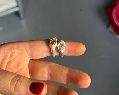 A hand with red nail polish holds two small rings against a gray indoor background. One ring is silver with a yellow gemstone, and the other is silver with a clear gemstone.